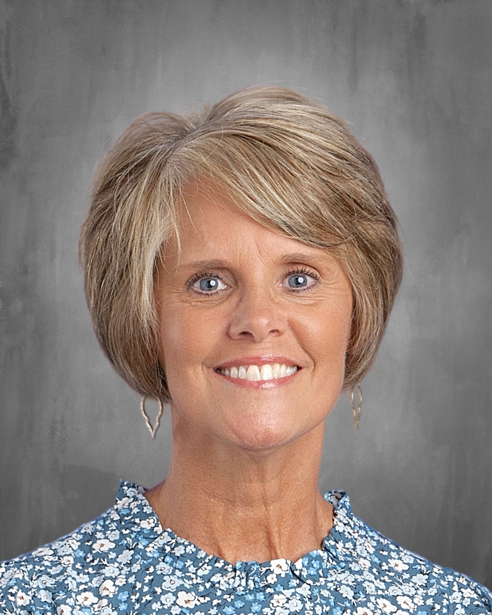 Woman with blonde hair, wearing glasses and a pink floral shirt smiles at the camera.