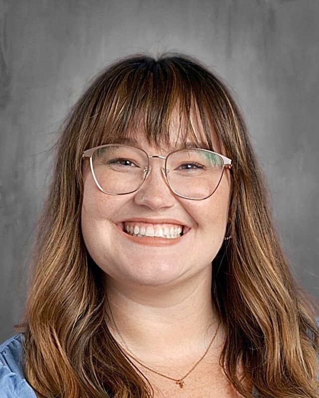 Woman with glasses smiling, wearing an orange top, against a gray backdrop.