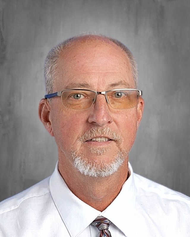 Man with glasses and goatee wearing a white shirt and patterned tie, posed against a gray backdrop.