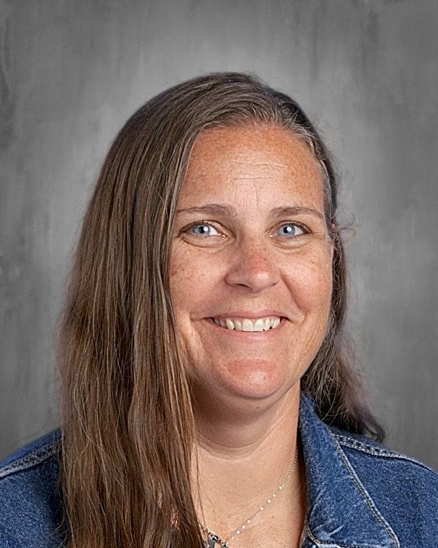 Woman with long brown hair, smiling, wearing a light blue top and necklace; gray background.