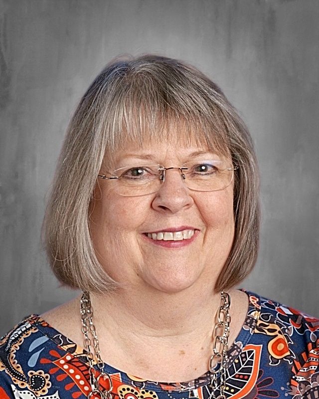 Smiling woman wearing glasses and a pearl necklace; headshot against a gray background.