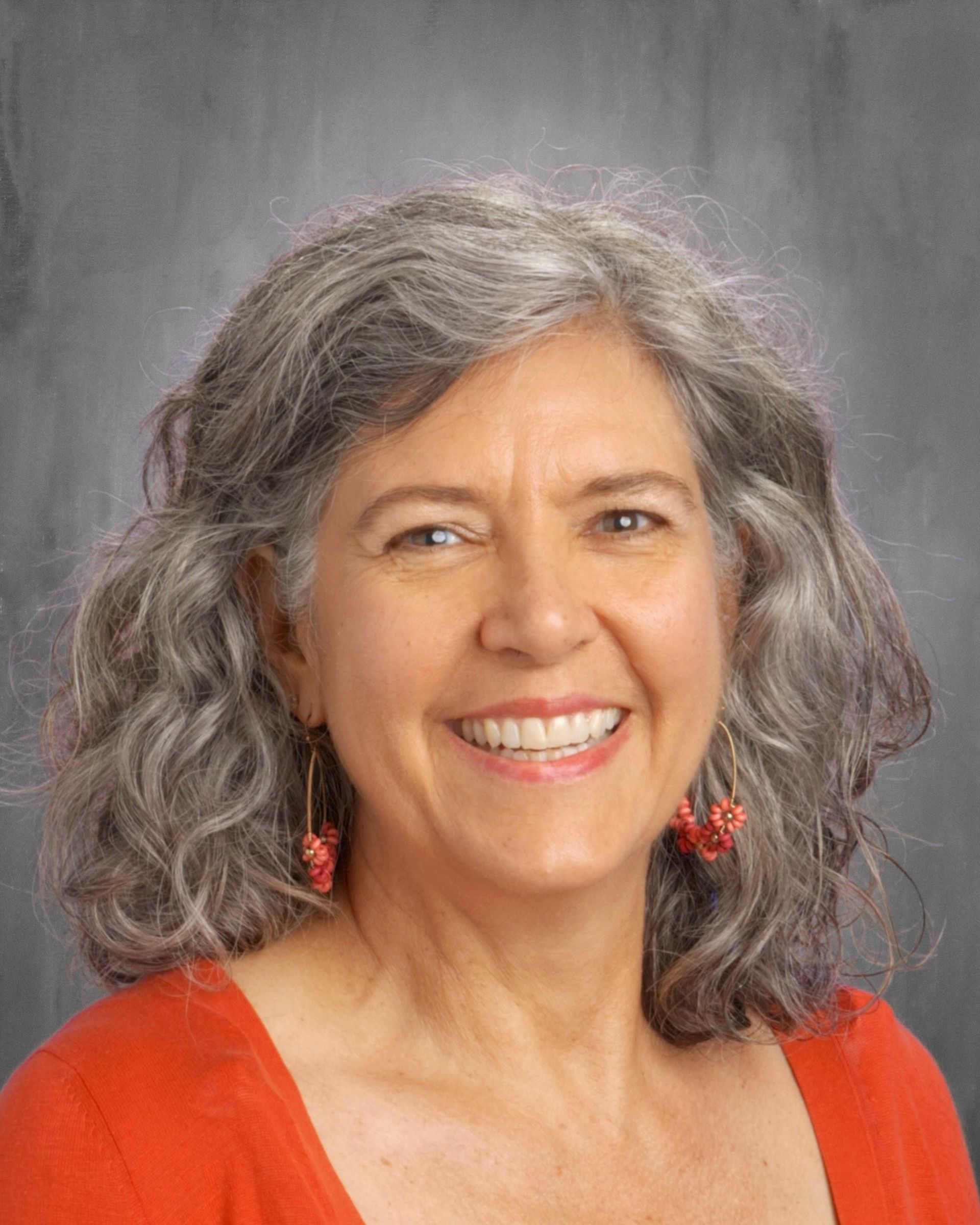 Smiling woman with gray curly hair, wearing orange top and earrings. Against a gray background.