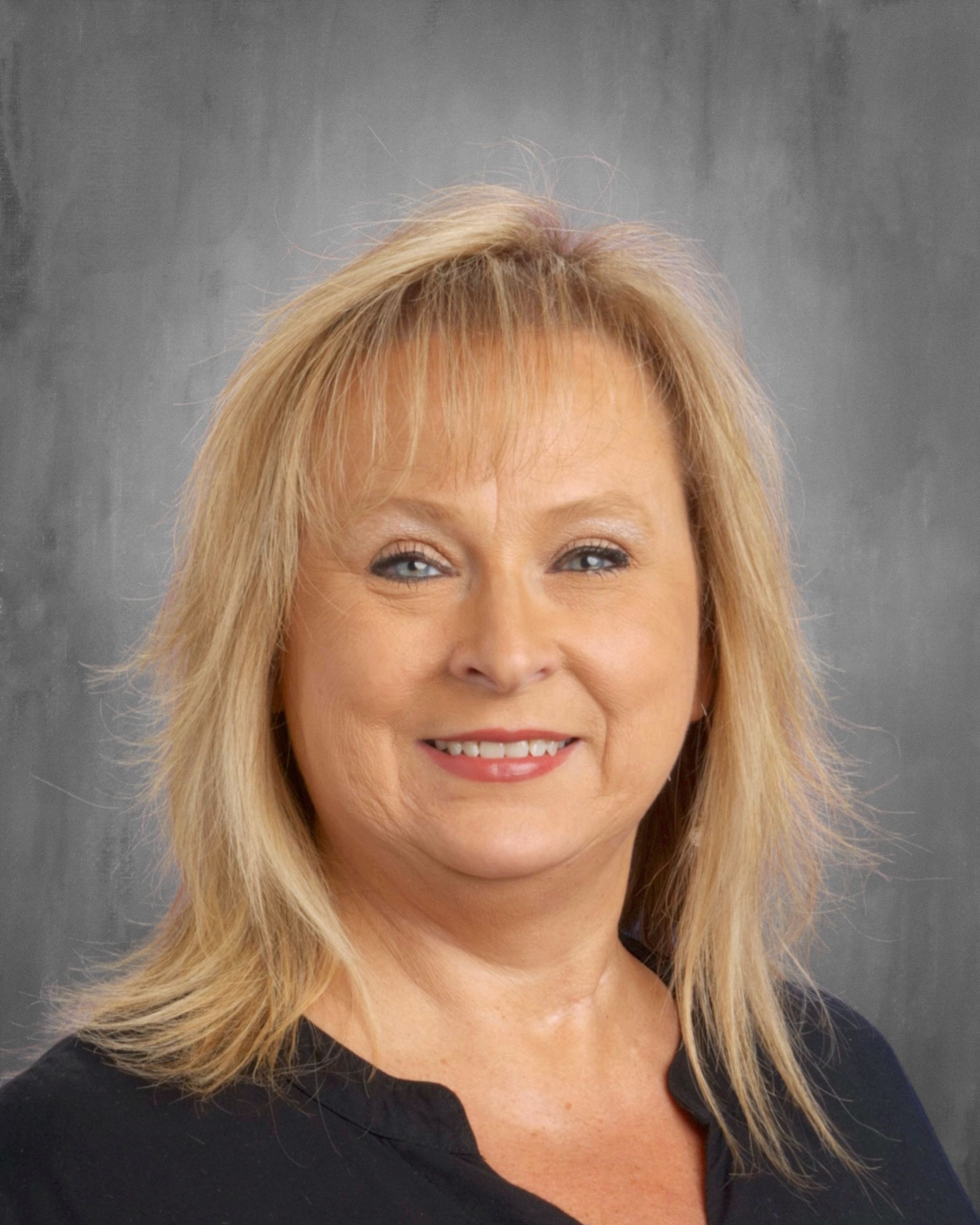 Blond woman with a slight smile, wearing a dark shirt, against a gray backdrop.