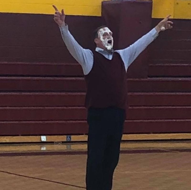 Man in maroon vest and shirt, face covered in cream, arms raised in celebration on a basketball court.