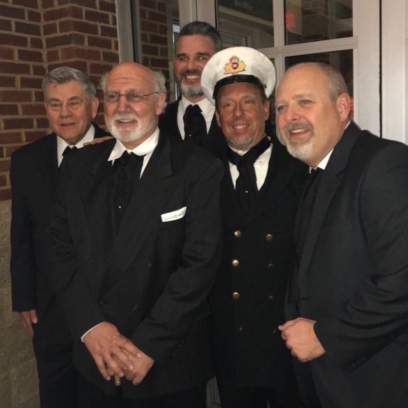 Five men in formal attire pose indoors. One wears a captain's hat and uniform, the others are in suits.