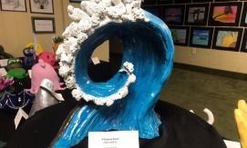 Blue wave sculpture with white foam at the crest, displayed on a table with art.