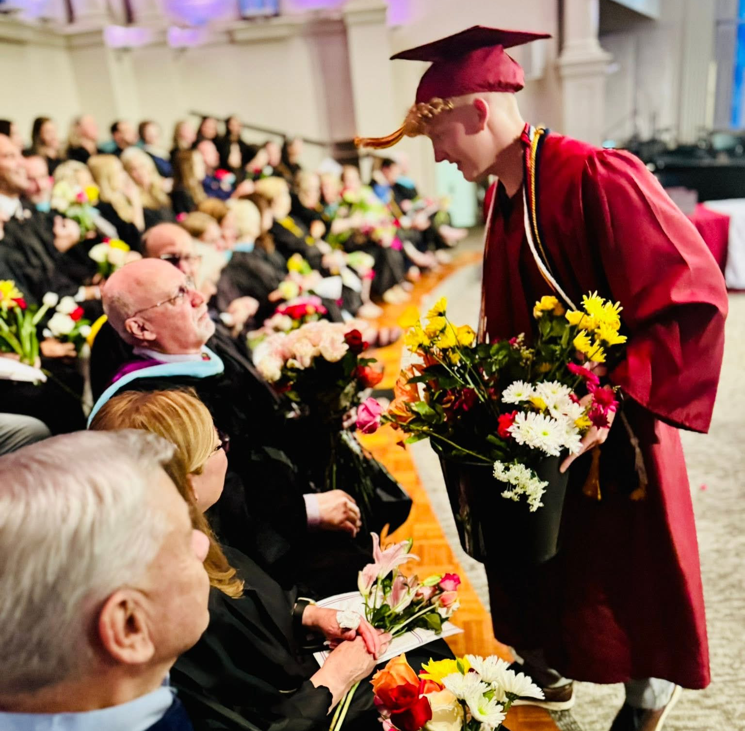 Graduate in maroon gown offers flowers to someone in the audience at graduation.
