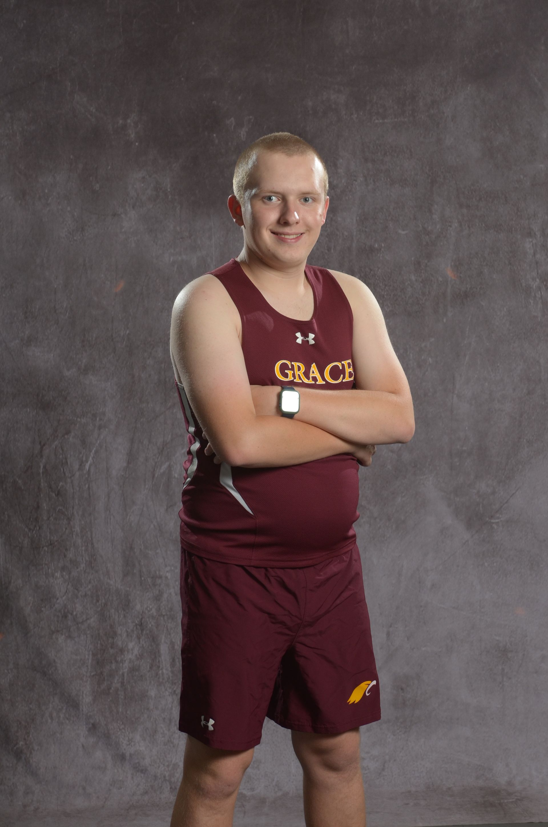 Teen track athlete standing on a track with arms crossed, wearing maroon uniform; outdoors.
