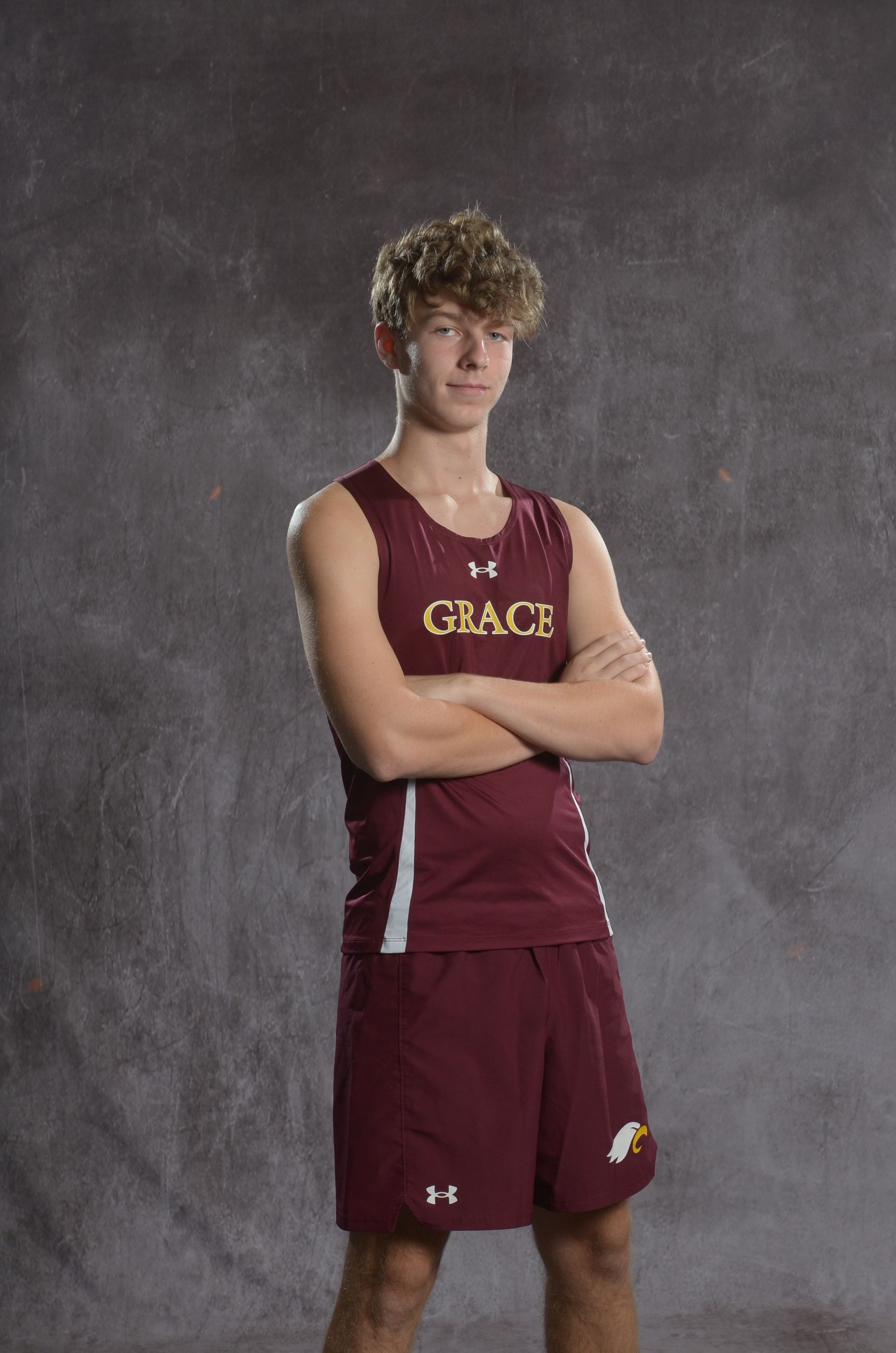Teenage track athlete standing on a field, arms crossed. Wearing maroon uniform, blue shoes, and smiling.
