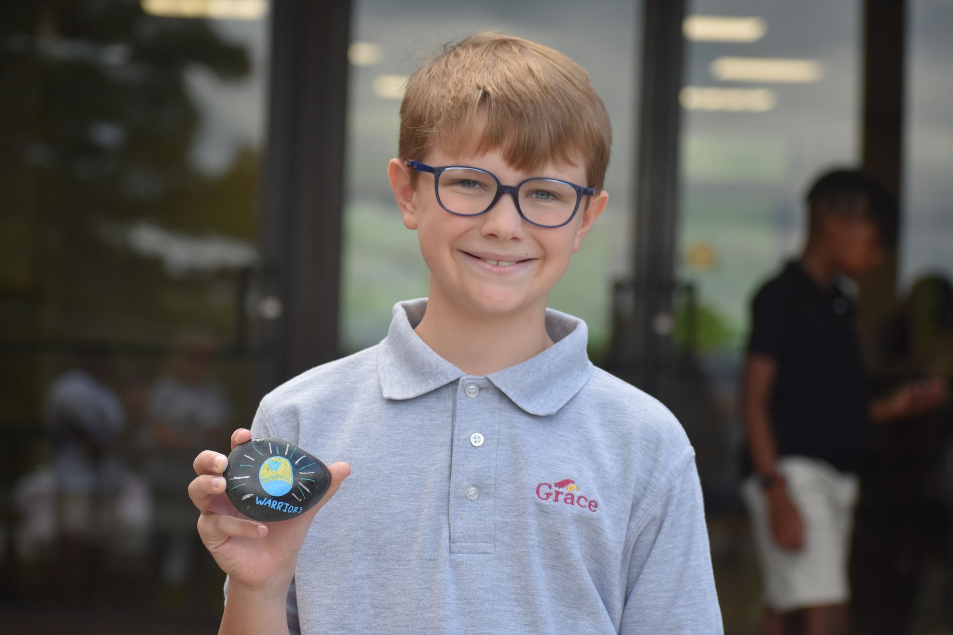 Boy in gray polo shirt holds a painted rock; smiling, outside a building.