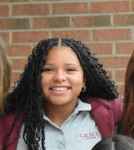 Smiling girl with long braids, wearing a gray polo shirt and burgundy jacket, stands in front of a brick wall.