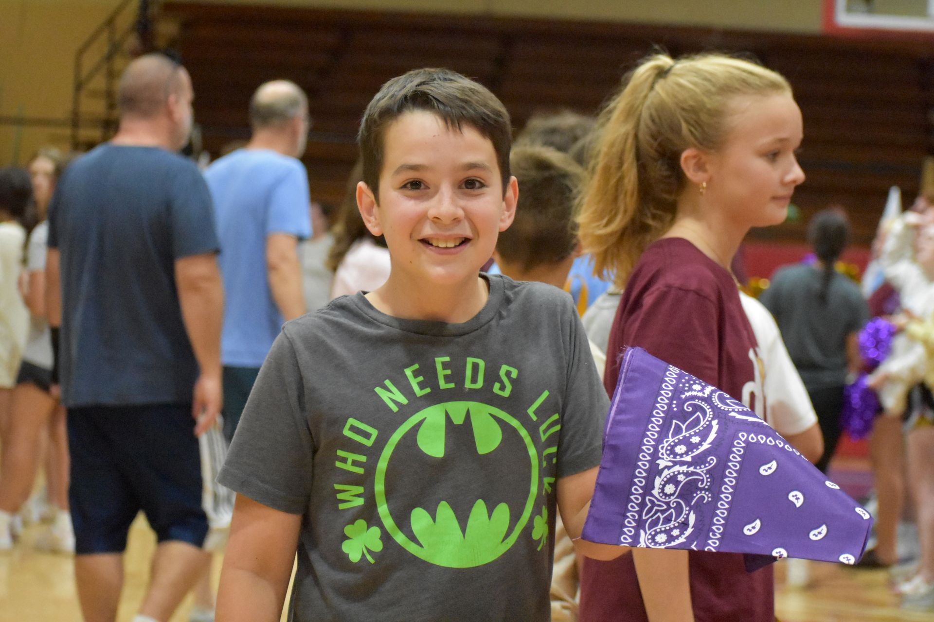 Boy wearing a Batman shirt smiles, holding a purple bandana in a gym. People in background.