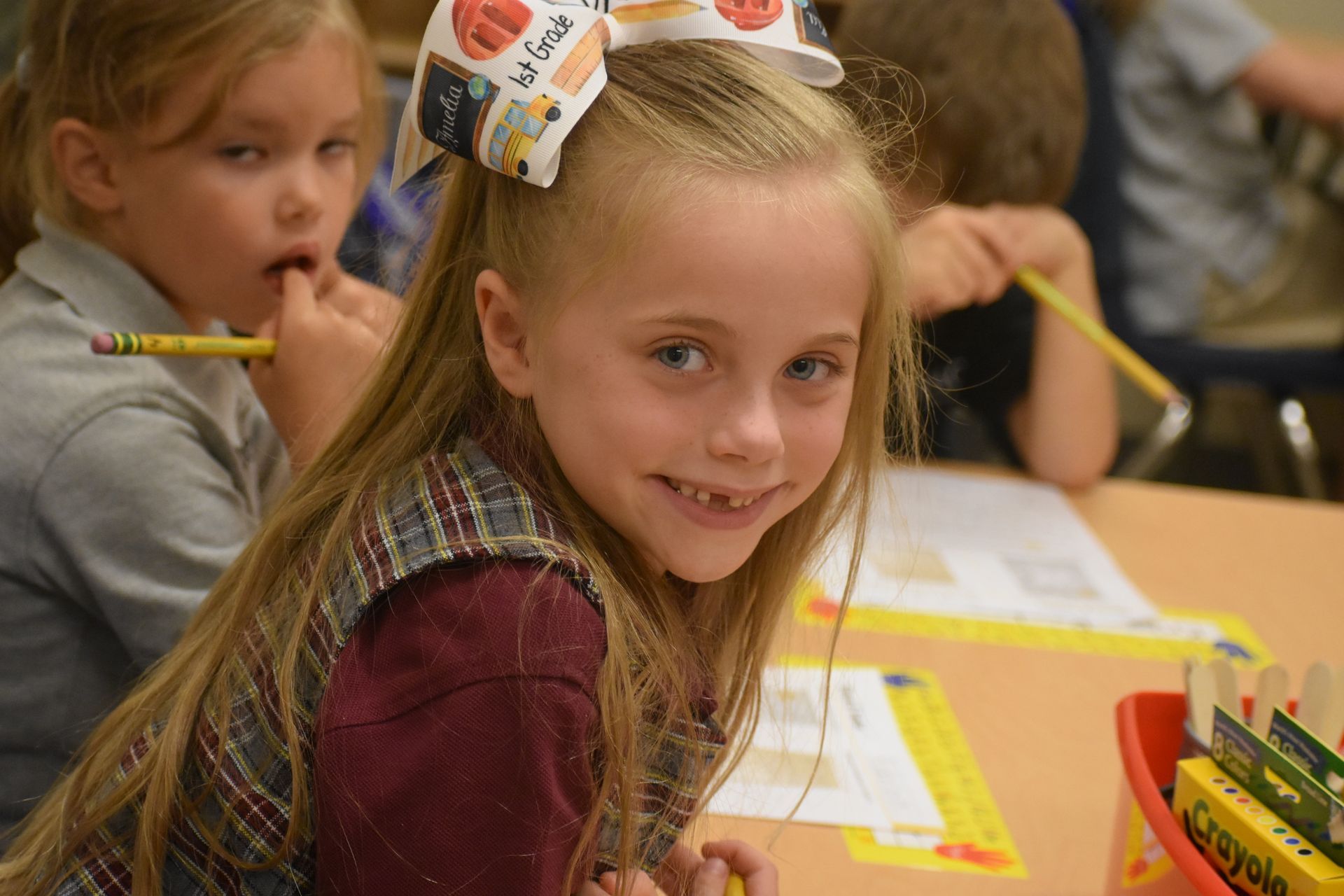 Young girl with missing tooth smiles at the camera in a classroom, bow in hair.