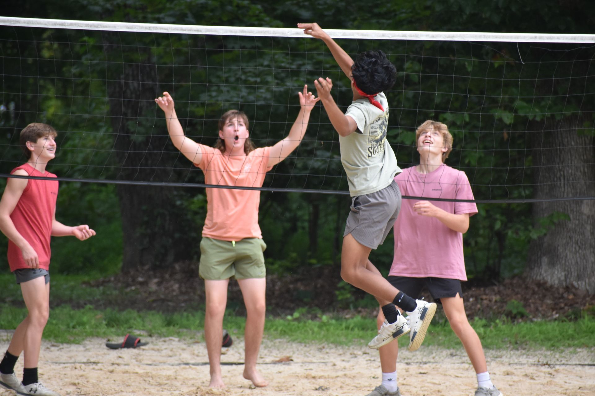 Four teens playing volleyball on a sand court; one jumps to spike, others watch or celebrate; outdoors, trees.