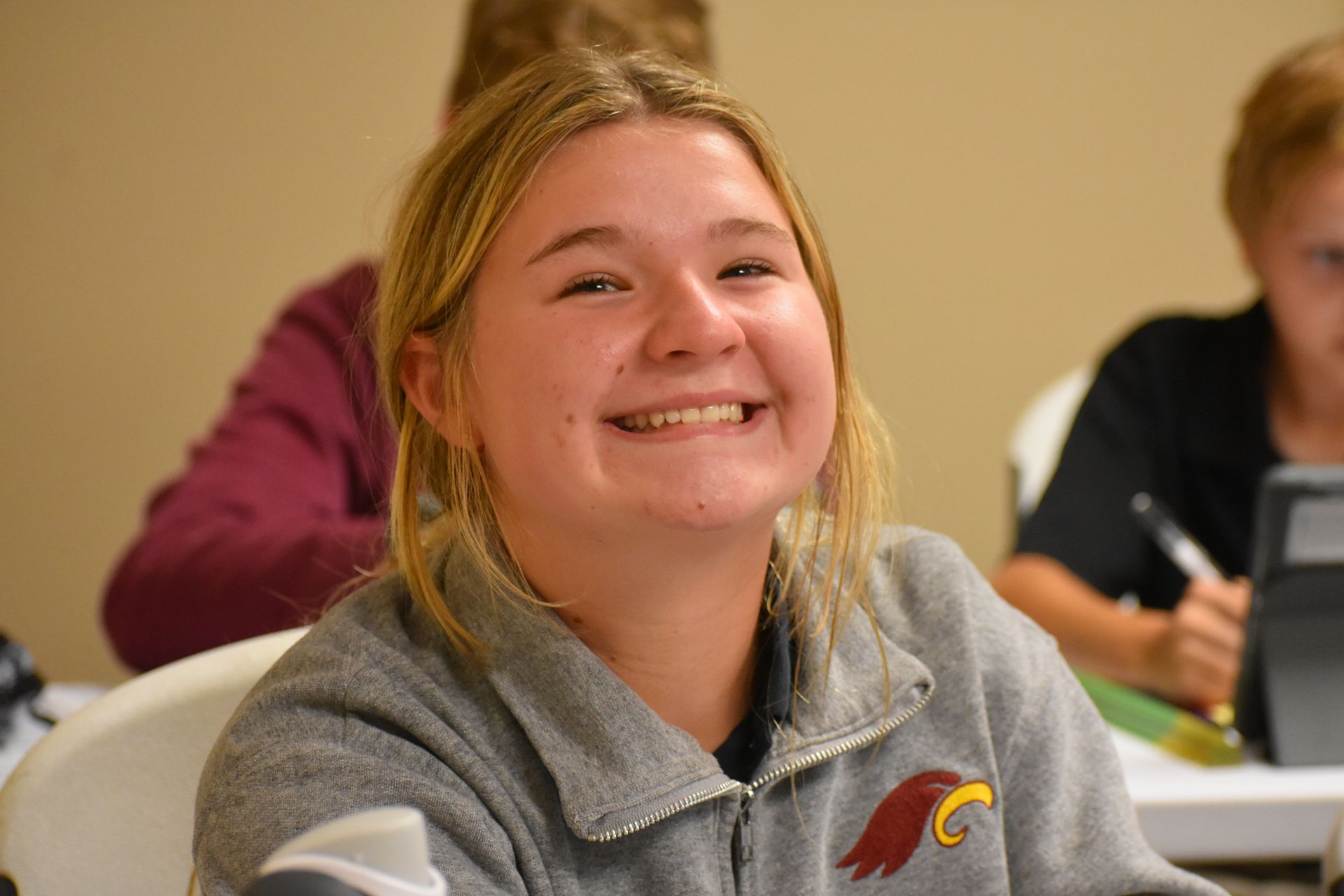 Blonde girl smiling broadly in a classroom setting, wearing a gray zip-up sweatshirt.