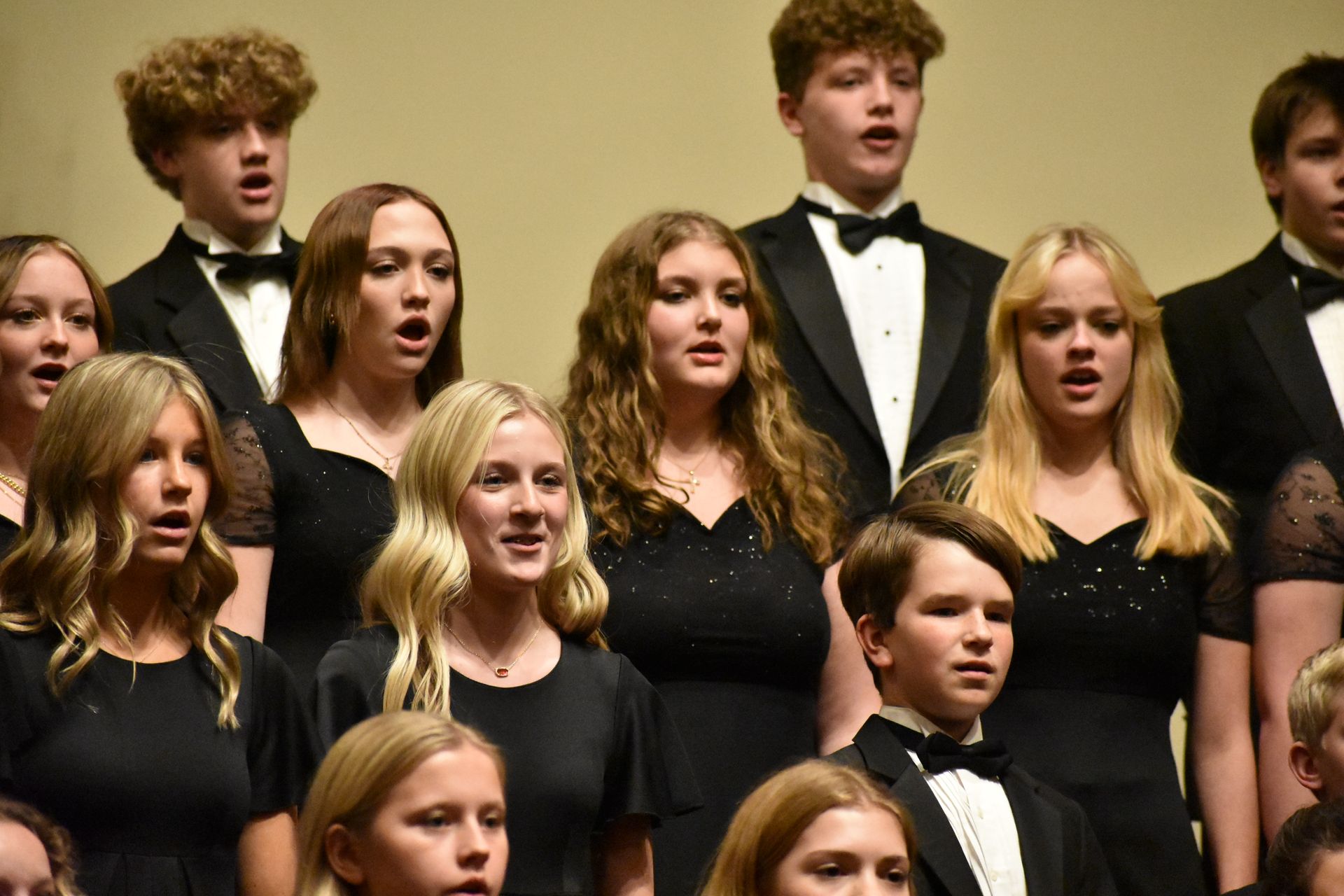 Choir performing in formal attire: black dresses and tuxedos. Concert hall setting, singing with focused expressions.