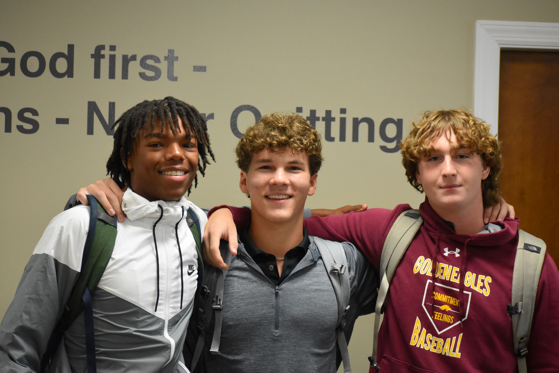 Three young men smiling, arms around each other, standing in a hallway. One has dreads, one has curly hair, and one has long hair.