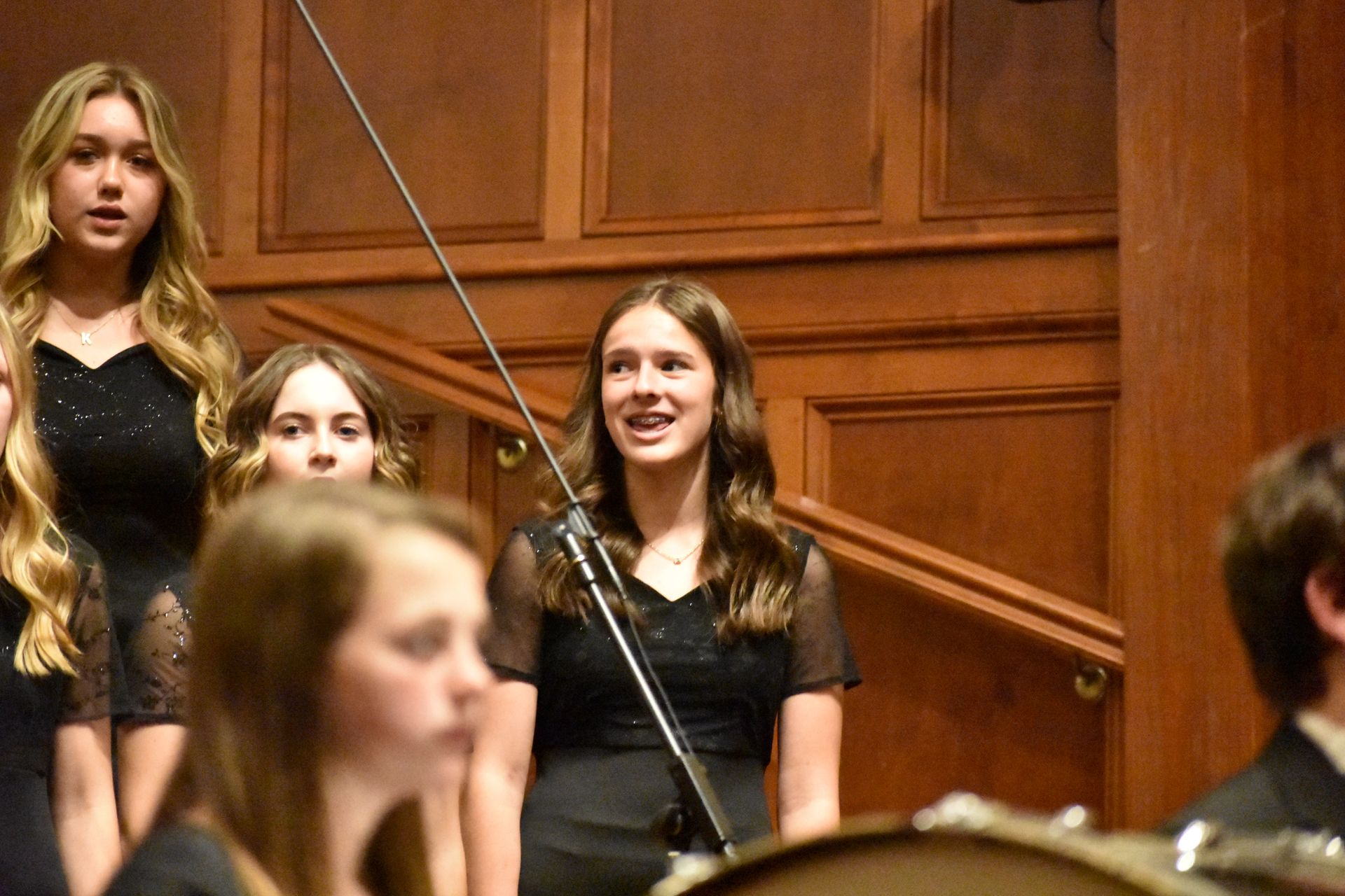 Choir in black dresses sing in a wooden-paneled room. A girl sings into a microphone.