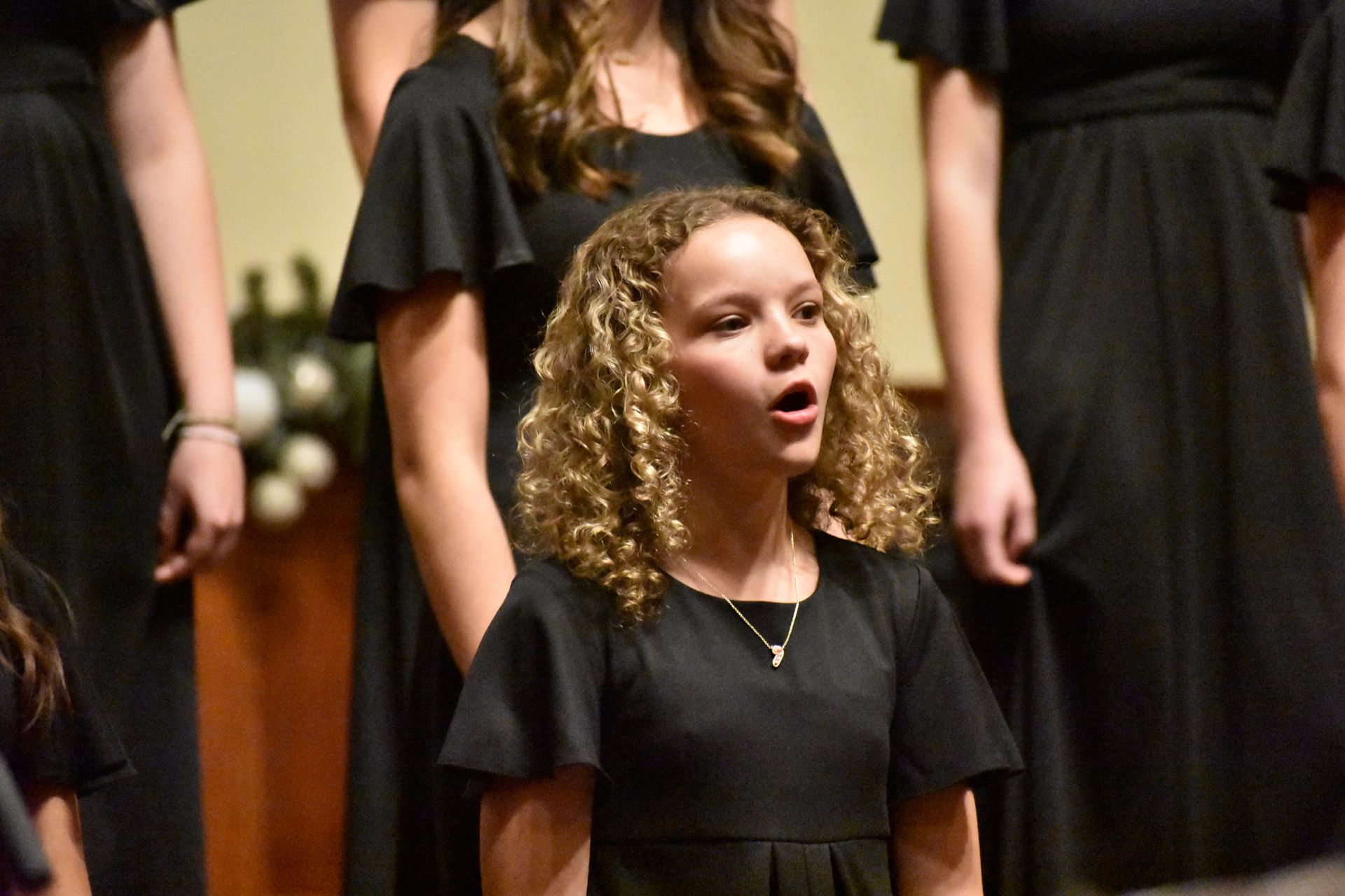 Choir girl singing, standing in front of other choir members. Wearing black dress, curly hair, lit setting.