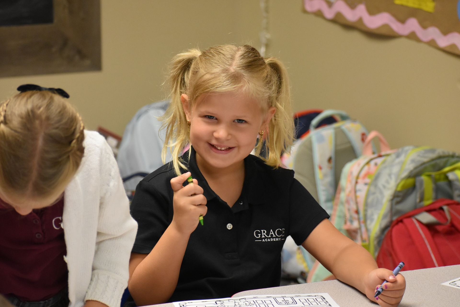 Girl with blonde pigtails smiles, holding a crayon at a desk, in a classroom setting.