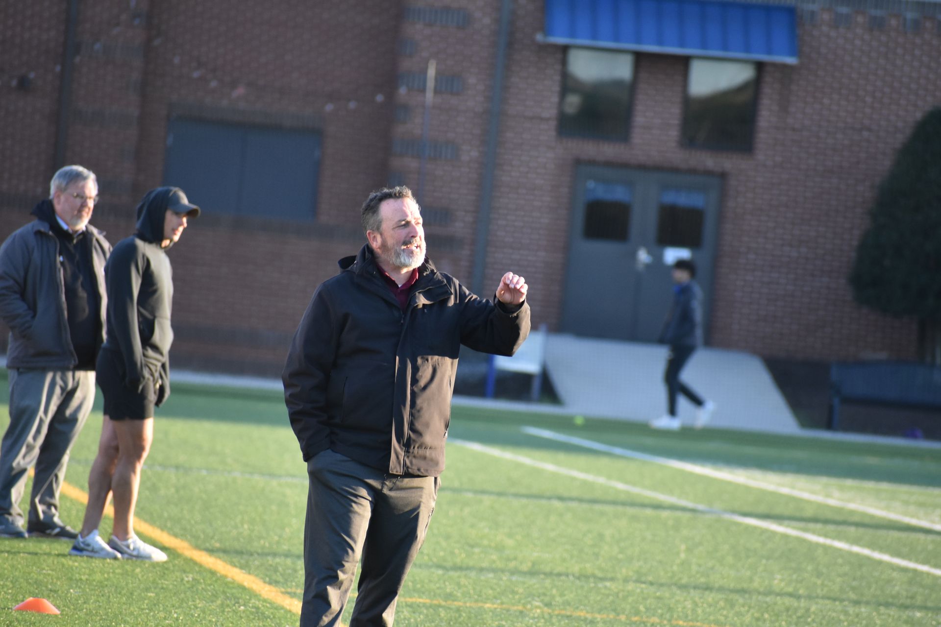 Man in black jacket gesturing on a green field with other people and a building in the background.