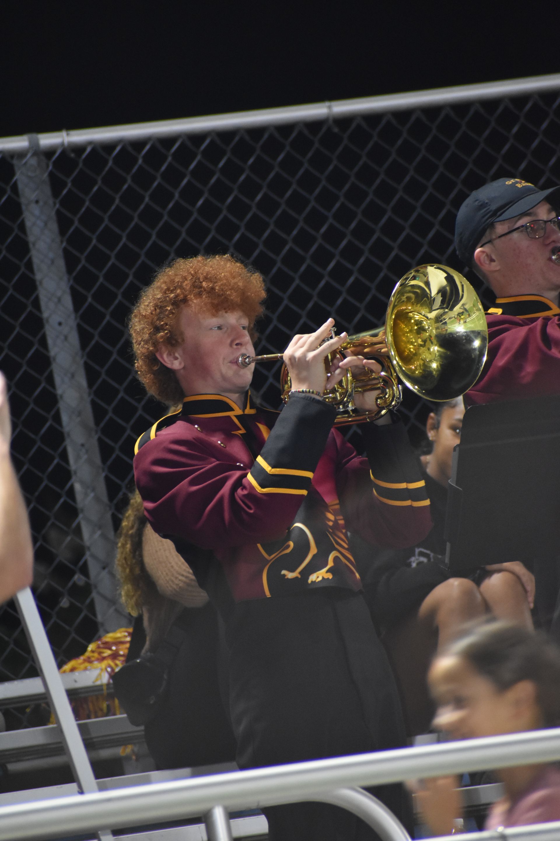 Young person with curly red hair plays a gold trumpet in a marching band, wearing a maroon uniform at a night game.