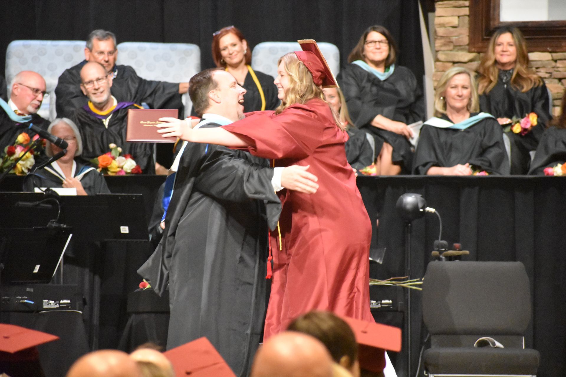 Man hugging graduate on stage at graduation ceremony.