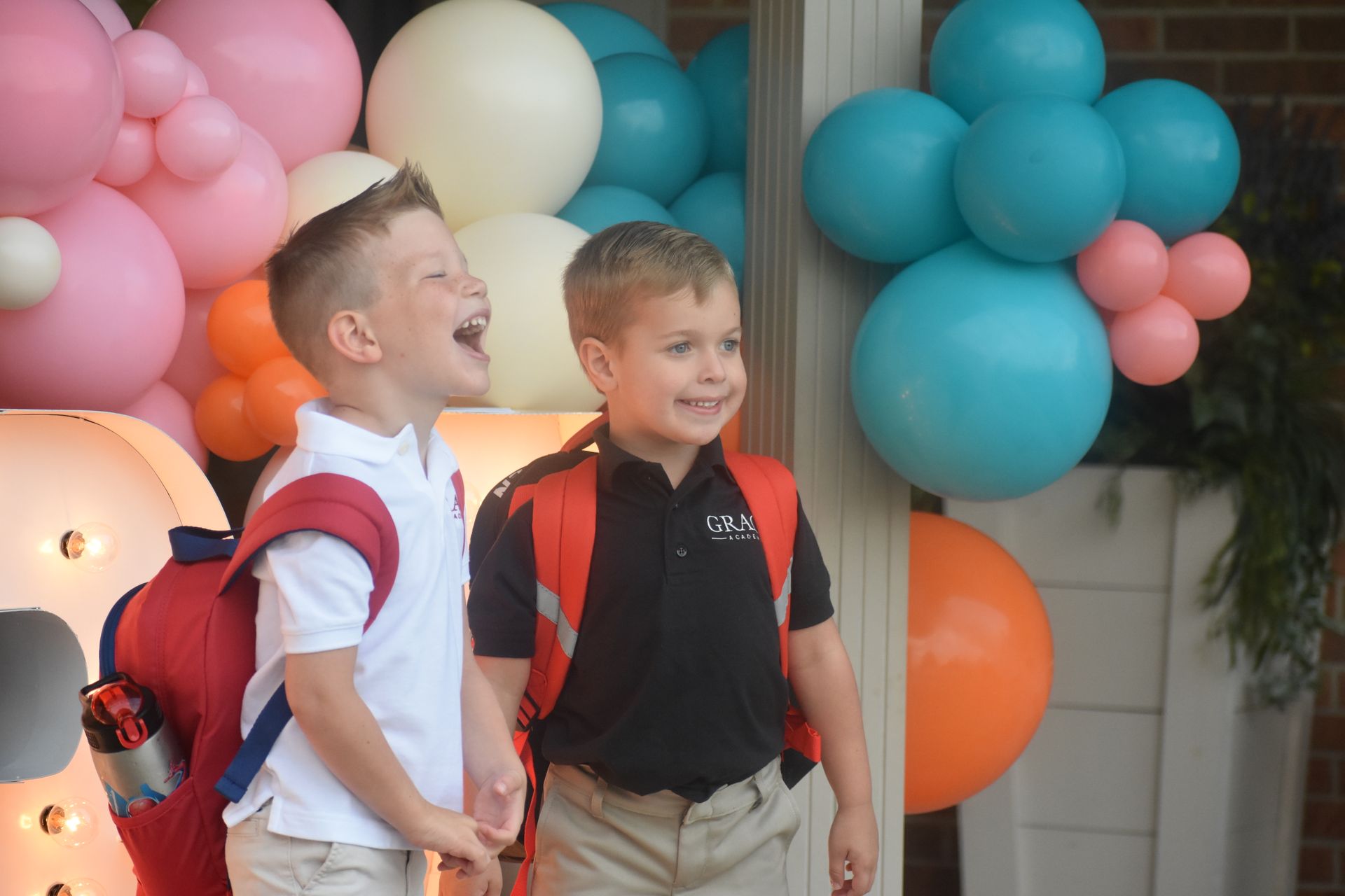 Two young boys, backpacks on, laughing in front of colorful balloons at an outdoor event.