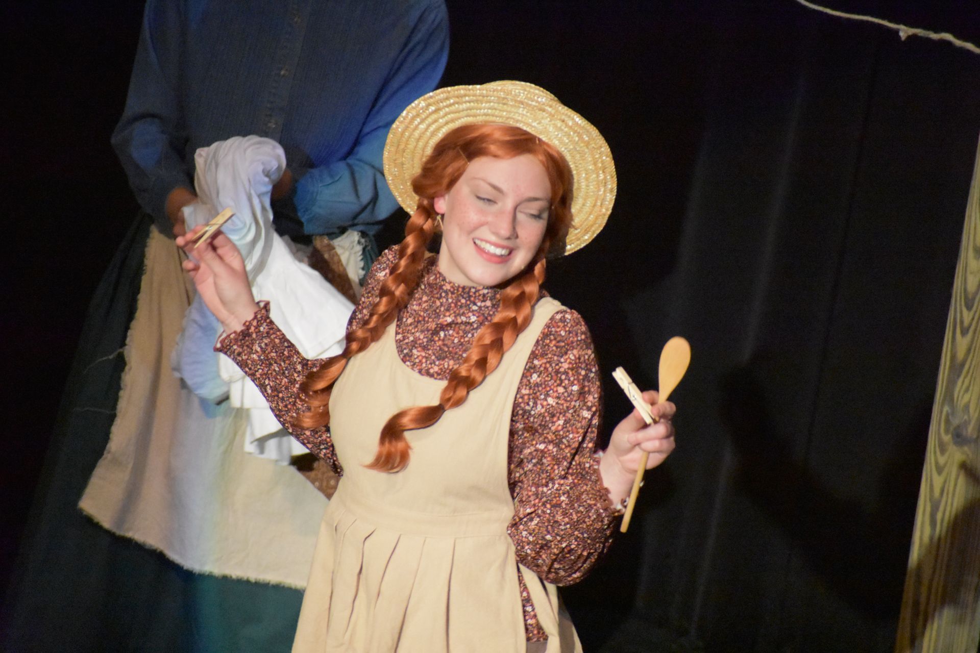 Woman with red braids, straw hat, and apron smiles, holding wooden spoons. Stage setting.