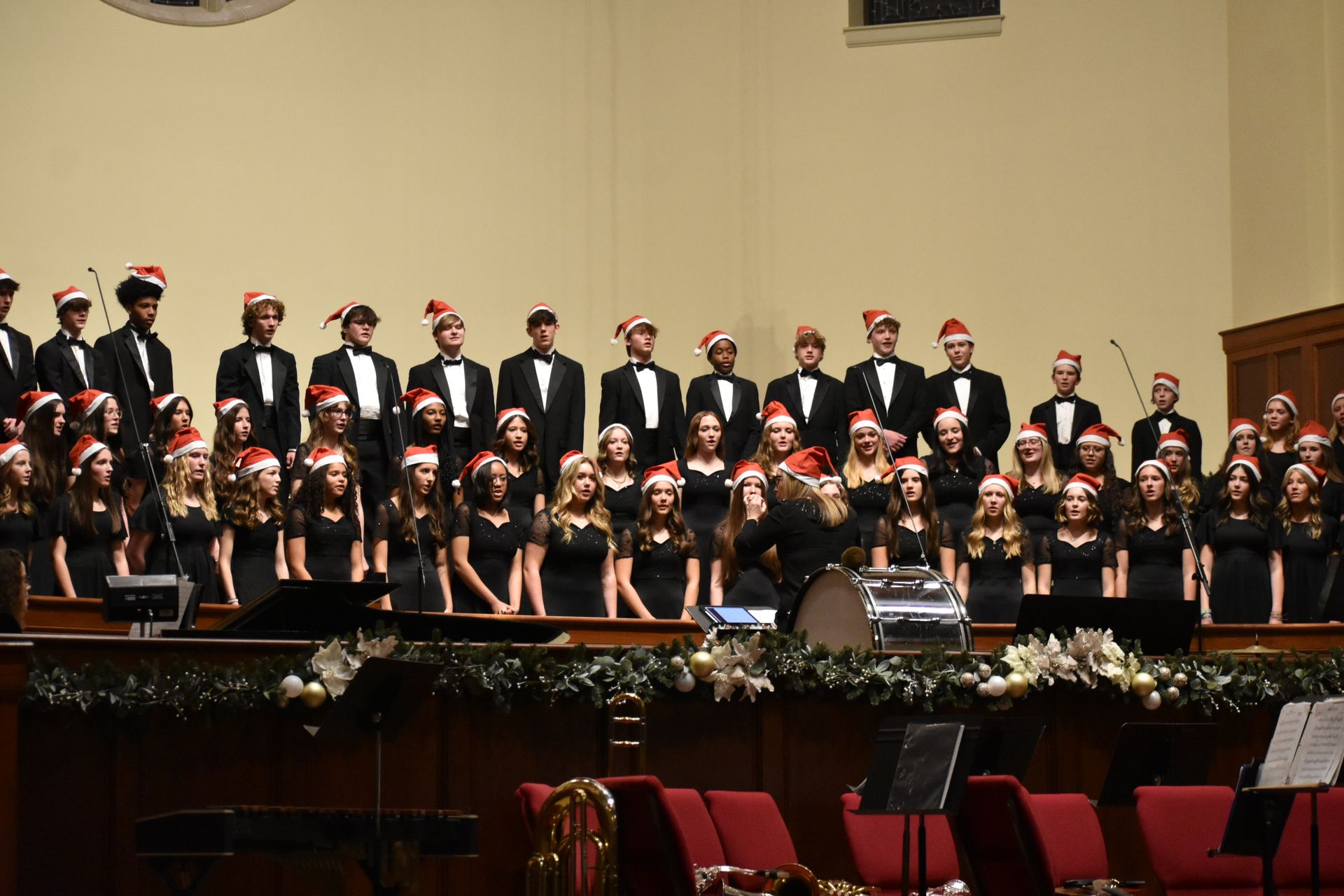 Choir of young people in black outfits and Santa hats singing on stage, decorated for the holidays.