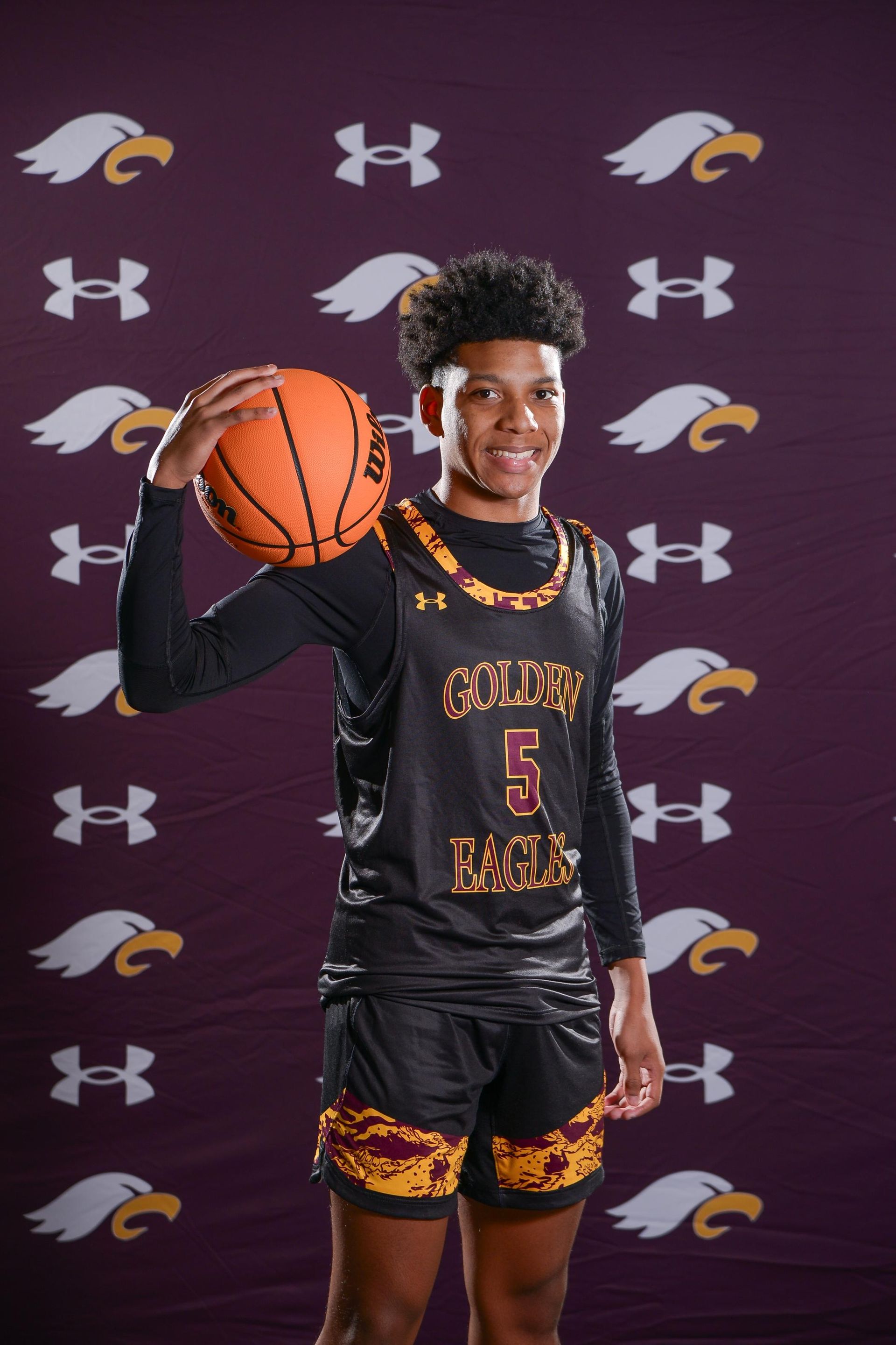 Basketball player in a black and gold jersey holding a Wilson ball, looking at the viewer.