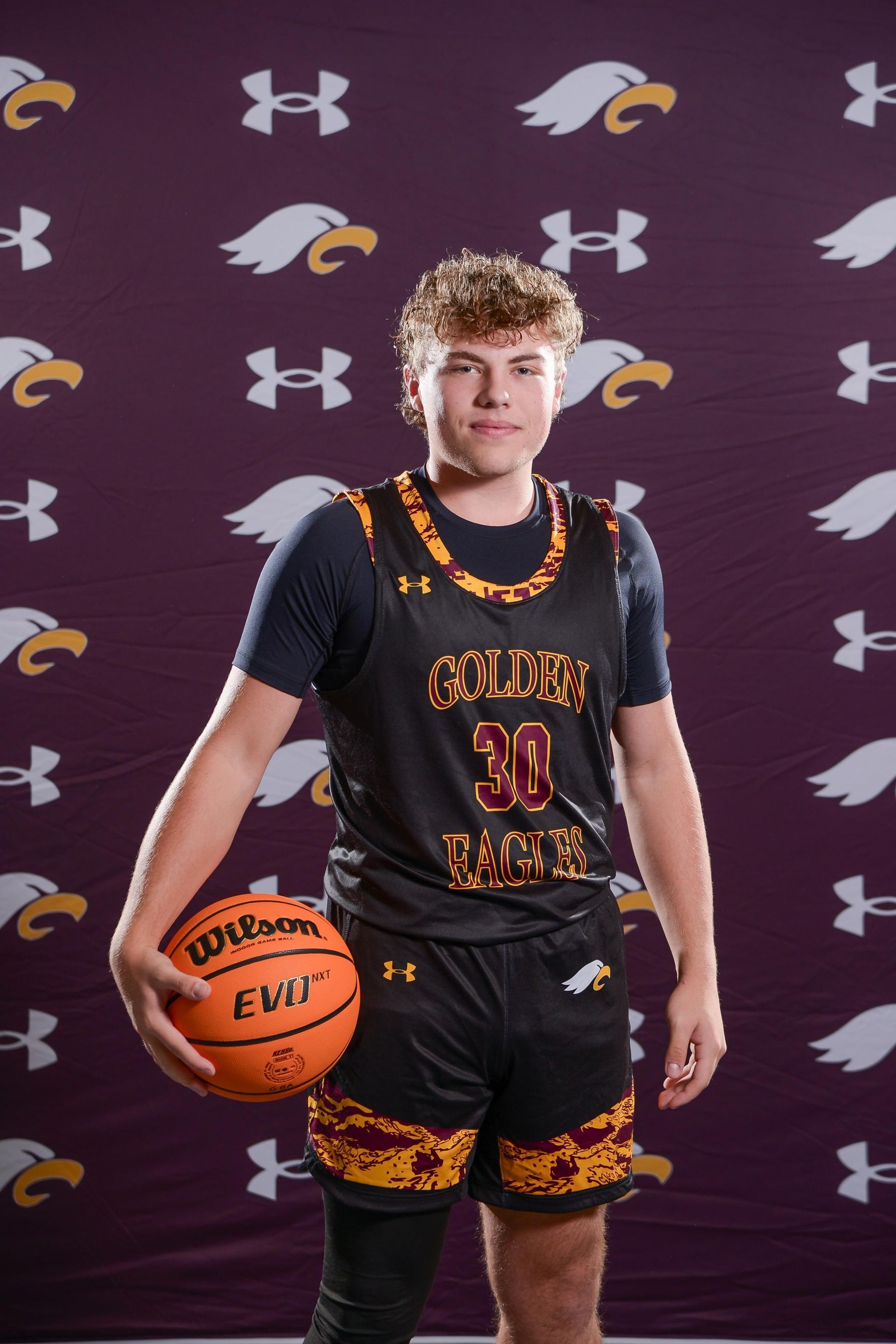 Basketball player wearing a black and gold jersey, holding a ball, in a dark setting.