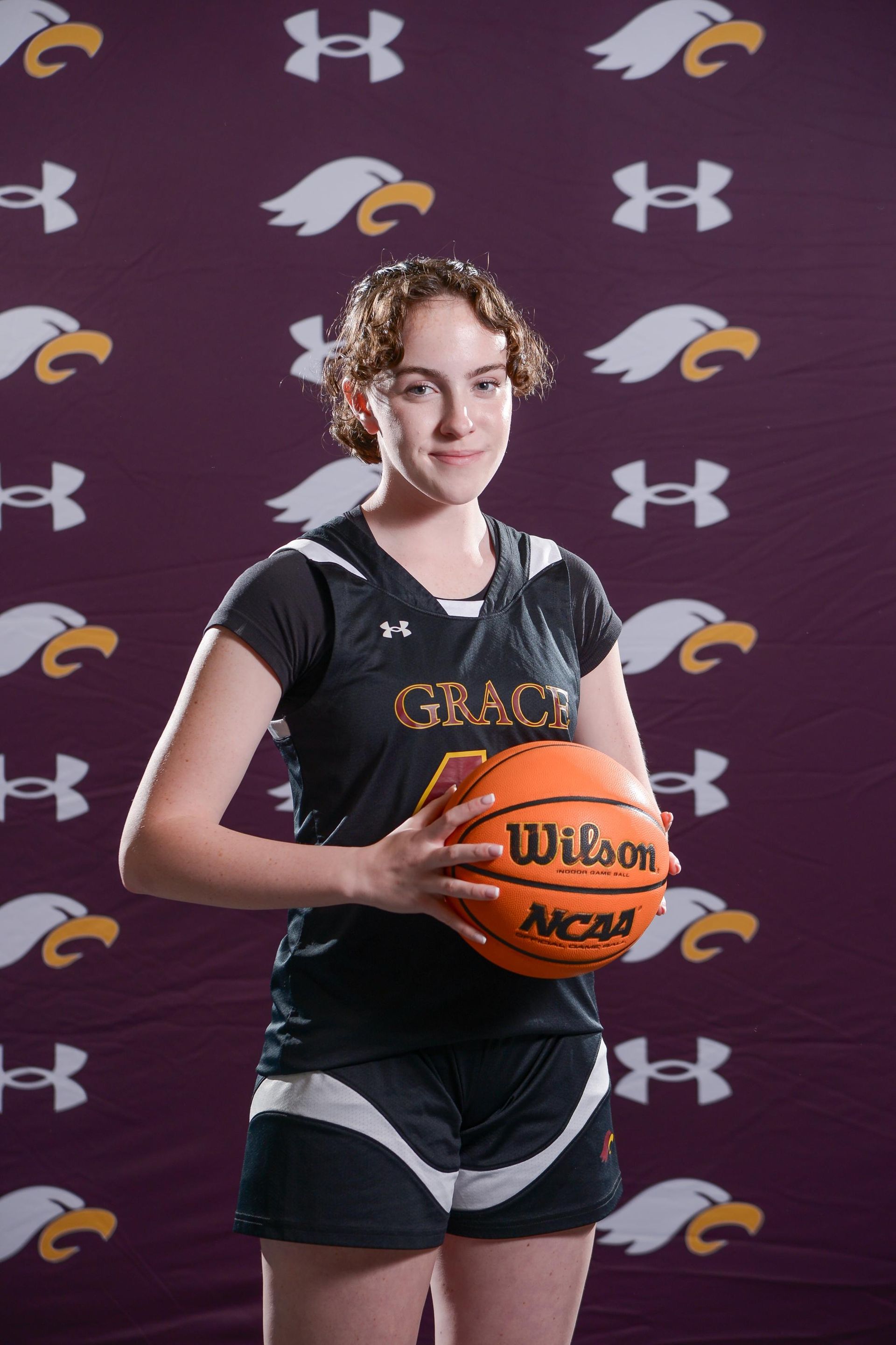A young woman with a shaved head in a Grace Academy basketball jersey holding a ball.