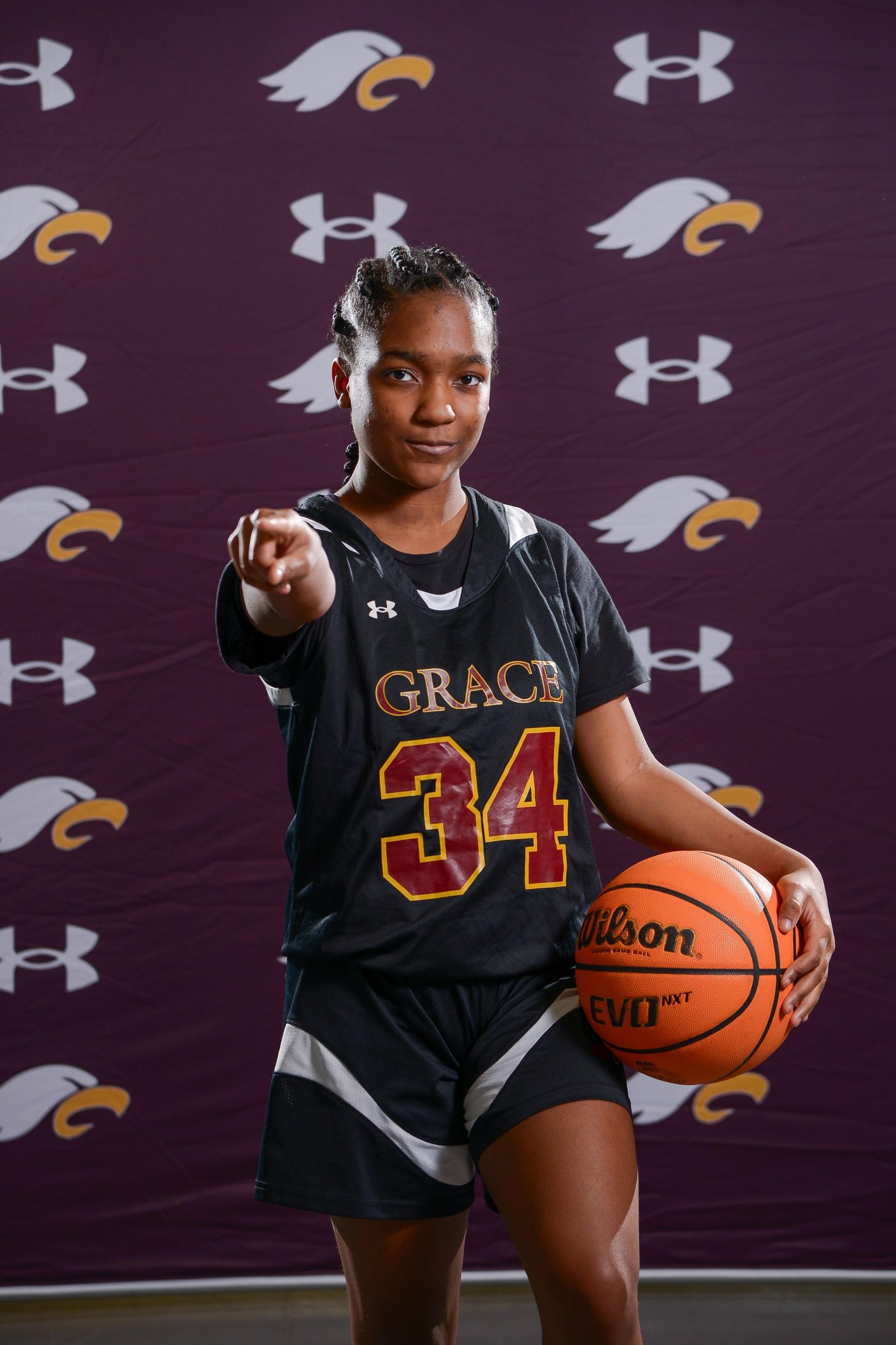 Young Black woman in maroon basketball uniform with number 34 holding ball, pointing at viewer.