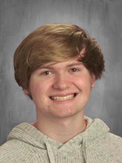 Smiling young man with light brown hair wearing a gray hoodie against a gray background.