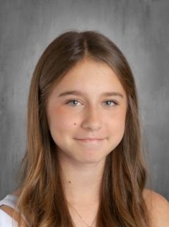 A young girl with light brown hair smiles at the camera against a gray backdrop.