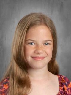 Blonde girl with a slight smile, wearing a floral shirt; gray background.