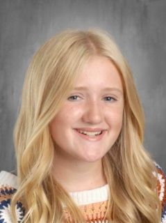 Blonde girl with a happy smile, wearing a patterned sweater, in front of a neutral background.