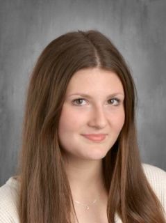 A young woman with long brown hair smiles, wearing a white sweater. Portrait on a gray backdrop.