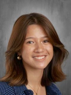 Smiling young woman with brown hair, wearing a blue top, against a gray background.