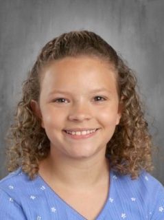 Young girl with curly brown hair smiles, wearing a blue shirt; studio portrait.