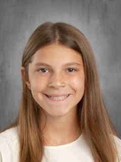 Girl with long brown hair smiles at the camera. Wearing a white top, plain gray background.