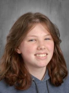 Teenager with reddish-brown hair, smiling, wearing a blue hooded sweatshirt, against a gray backdrop.