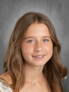 Girl with long brown hair, braces, smiles, wearing a necklace. Gray background.