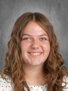 Young woman with long wavy brown hair smiles, wearing a white patterned shirt, in front of a gray backdrop.