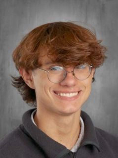 Young person with reddish-brown hair and glasses smiling at the camera, gray background.