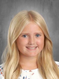 Blonde girl with blue eyes smiles, light-skinned, wearing a light top, headshot, neutral background.