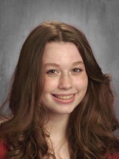 Young woman with long brown hair smiling, looking at the camera. Red shirt, neutral background.
