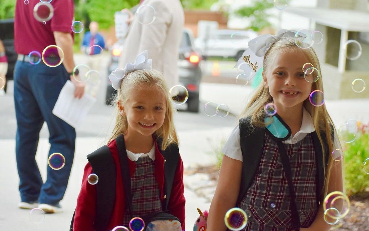 Two young girls, smiling, in school uniforms with bows, surrounded by bubbles.