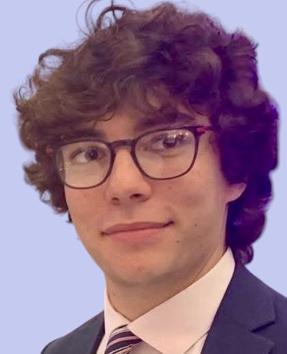 Young man with curly brown hair and glasses, wearing a suit, smiles.